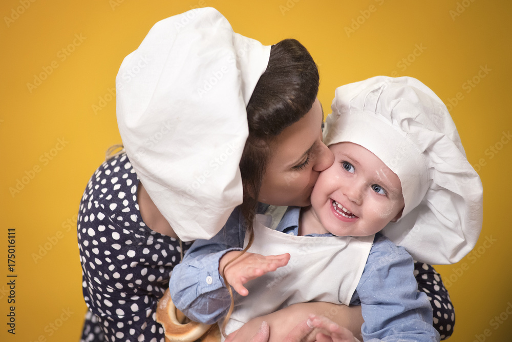 Mother kisses her smiling son on the cheek in the hat chef on a yellow background Mother kisses her smiling son on the cheek in the hat chef on a yellow background