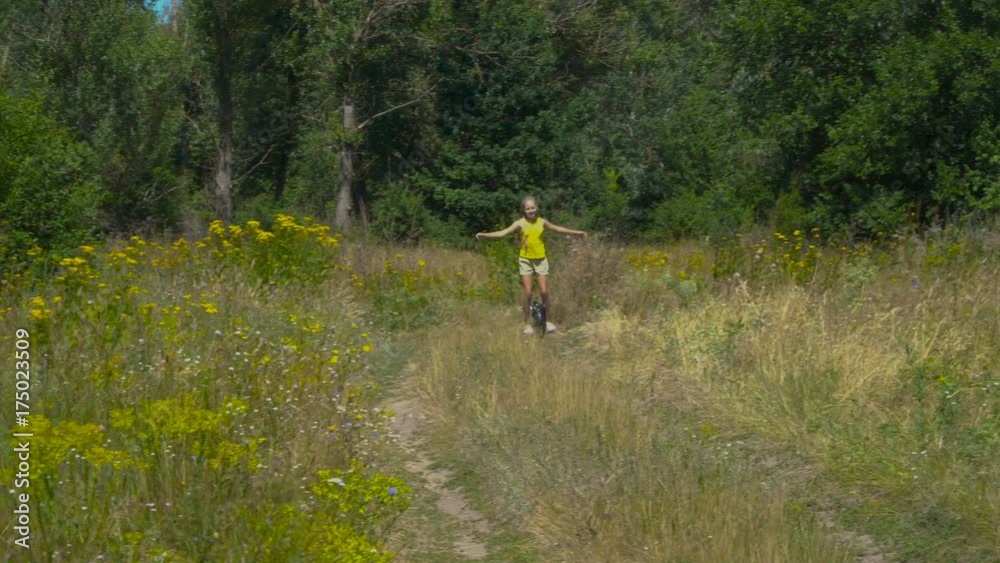 The girl is riding on a monocycle along the field road. The child is wearing a yellow jersey. She is happy and smiling. The girl is riding on a monocycle along the field road. The child is wearing a yellow jersey. She is happy and smiling.