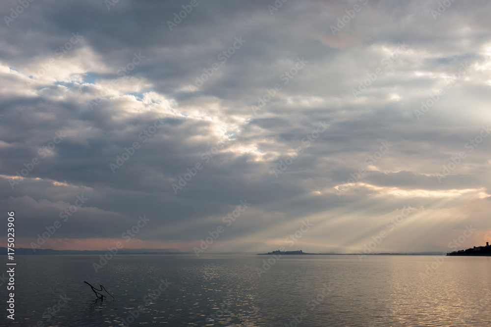 Sun rays coming out through the clouds over an island on a lake, with some branches in the foreground