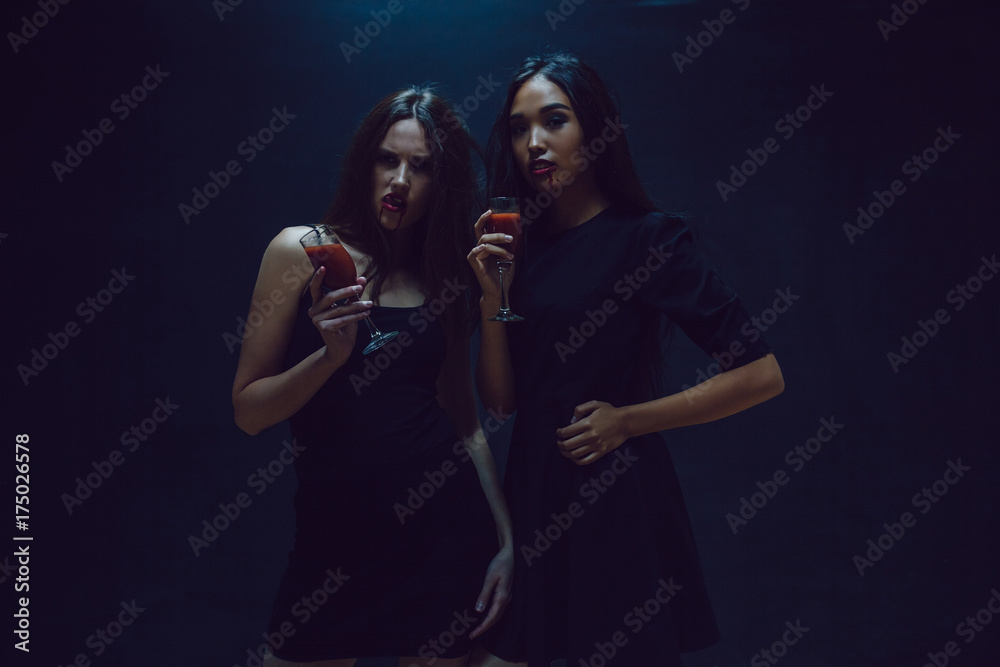 Attractive girls in the image of vampires hold glasses with blood. Halloween. Attractive girls in the image of vampires hold glasses with blood. Halloween.