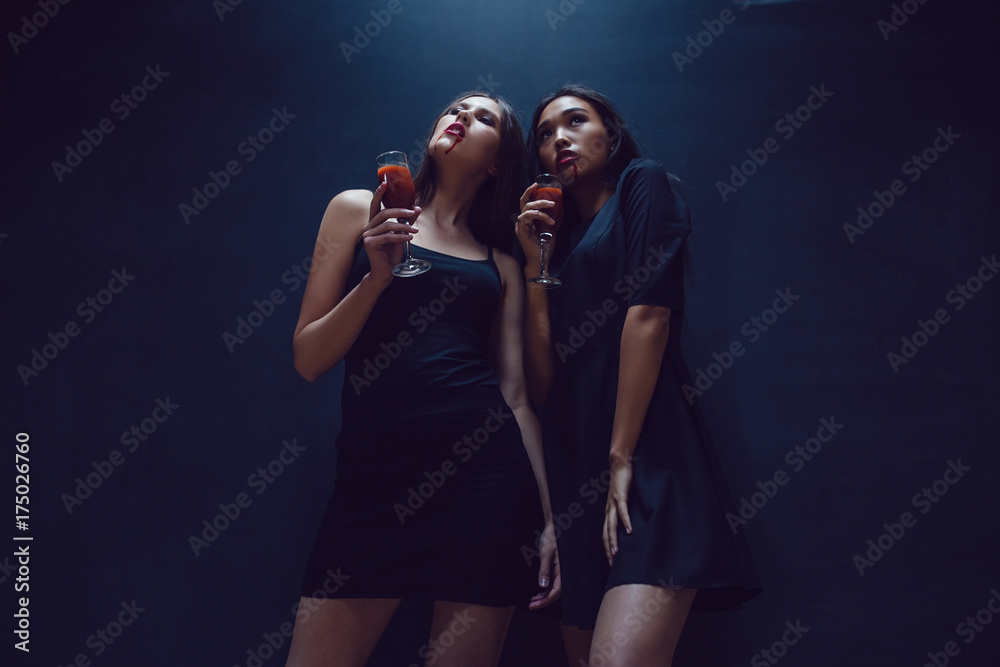Attractive girls in the image of vampires hold glasses with blood. Halloween. Attractive girls in the image of vampires hold glasses with blood. Halloween.