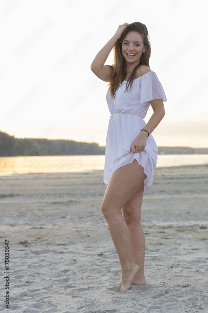 Young beautiful girl in white dress posing on the sandy beach with sunset and river on the background Young beautiful girl in white dress posing on the sandy beach with sunset and river on the background