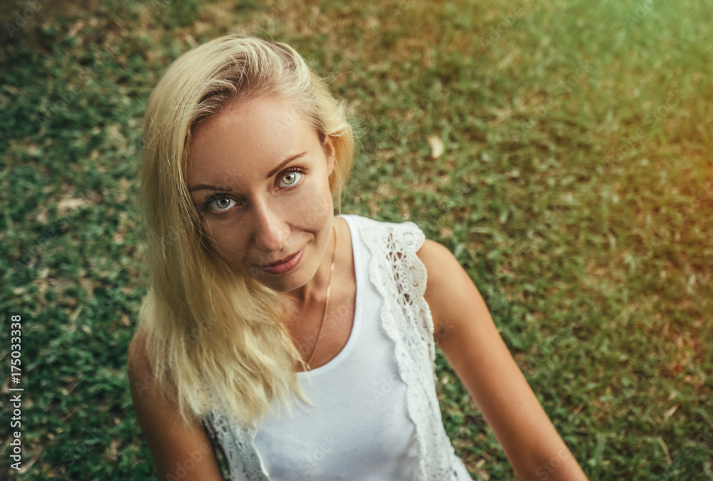 Beautiful young blond woman sits on the green grass and looks at the camera with beautiful gray eyes. Modern lifestyle and relaxation concepts Beautiful young blond woman sits on the green grass and looks at the camera with beautiful gray eyes. Modern lifestyle and relaxation concepts