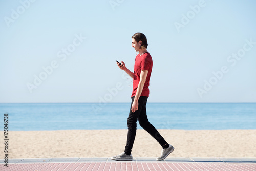 Full body side of handsome man walking by sea with smart phone