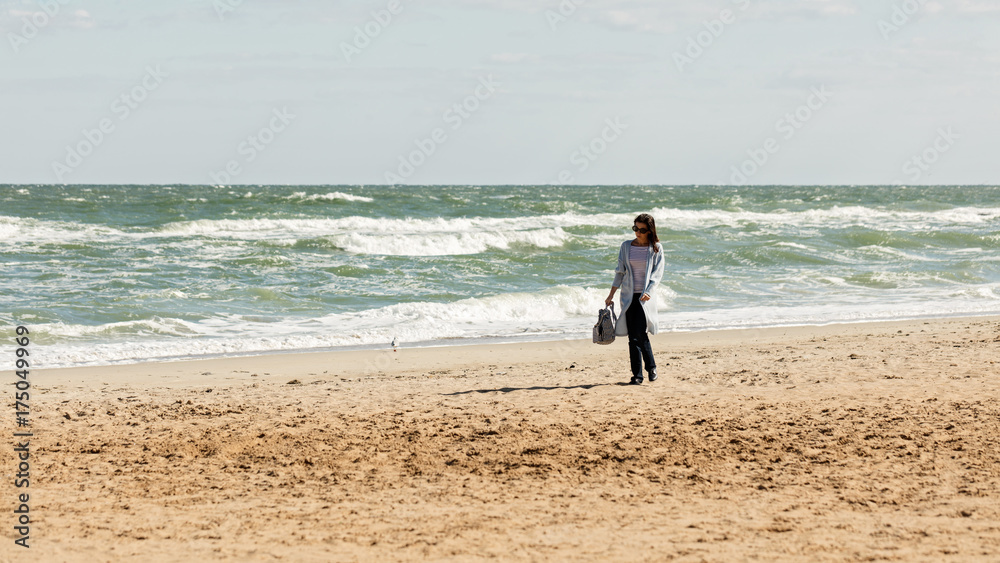 Young woman (brunette) in a light blue cardigan and navy jeans, with a striped backpack, walks along the beach. Young woman (brunette) in a light blue cardigan and navy jeans, with a striped backpack, walks along the beach.