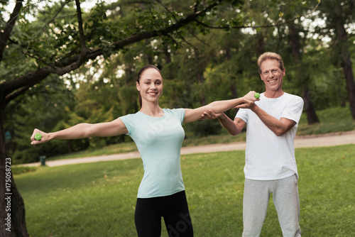 Girl doing exercises with dumbbells in the park. A man helps her. They are smiling