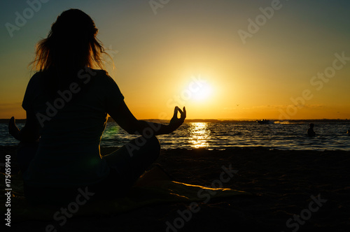 Woman practices yoga and meditates in the lotus position on the beach at sunset, rear view