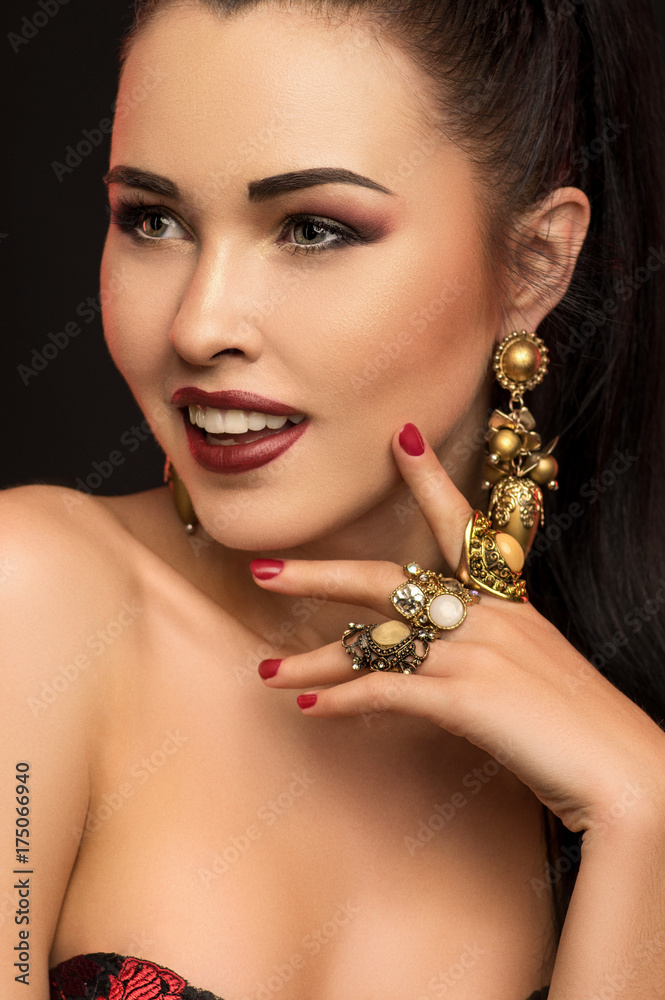 Fashion studio portrait of young brunette model with stylish earrings touching face, dark background