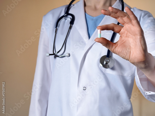 A female doctor with a stethoscope on a beige background holds a pill.