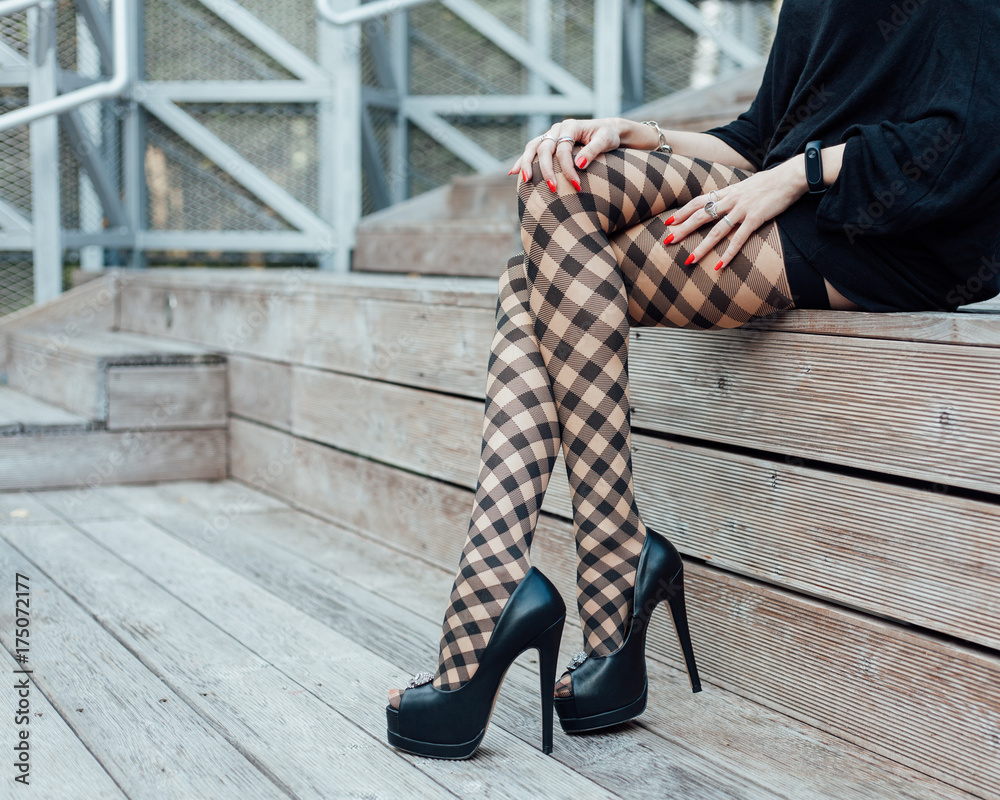 Foto de A sexy girl with long legs in fashionable, fishnet stockings, a  short dress and black high-heeled shoes sits in summer on wooden steps,  throwing a leg on her leg. Fashion.