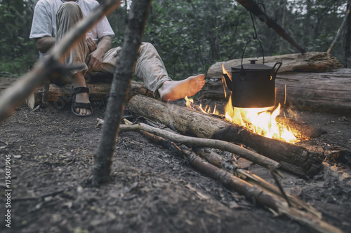 Man sits and heats his feet near the camp fire, over which the kettle hangs
