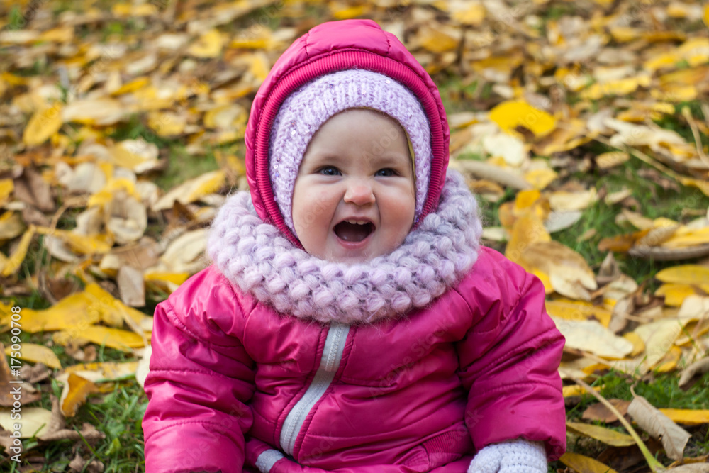 Toddler girl in pink jumpsuit and white mittens sitting on leaves in autumn park and smiling Toddler girl in pink jumpsuit and white mittens sitting on leaves in autumn park and smiling