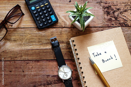 Wood Desktop work space have brown glasses,calculator,pot plant ,black watch,pen,notebook,and note pad with "Take a break"message.For Work life balance concept,Happy work place,Have copy space.