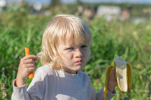 Blond Slavic Baby boy eats carrot and banana sitting on grass during outing at a summertime - healthy feeding