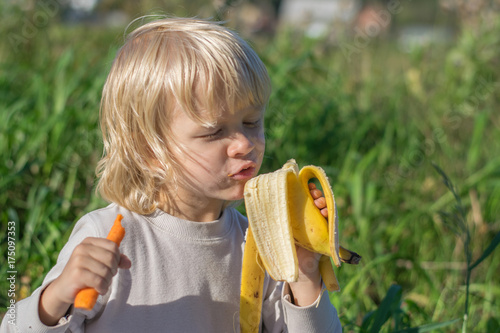 Obraz na płótnie Blond Slavic Baby boy eats carrot and banana sitting on grass during outing at a summertime - healthy feeding w salonie