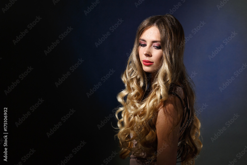 insanely beautiful blonde girl with blue eyes, pouting lips, curly hair,  with makeup, in a pink top, close-up stands on a blue background, turning  to the side and looking down Stock Photo |