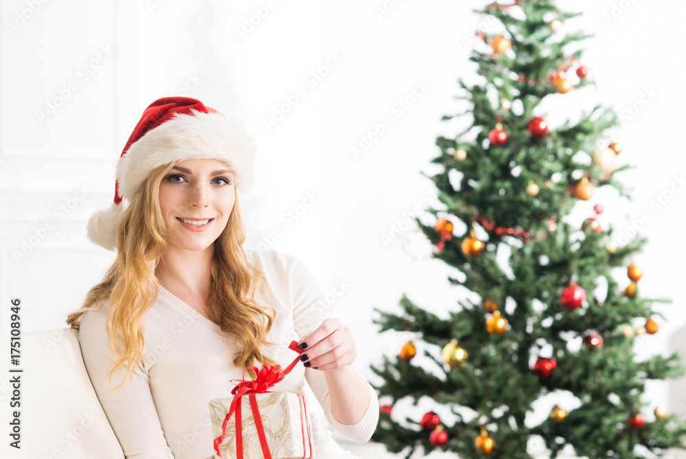 Beautiful blond girl celebrating Christmas at home. Young woman with xmas present. Tree on a background. Holidays and celebration concept.
