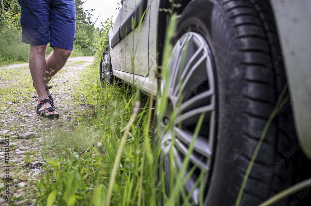 Man wearing shorts and sandals suitable to the car that is parked on the roadside near the forest, summer day.