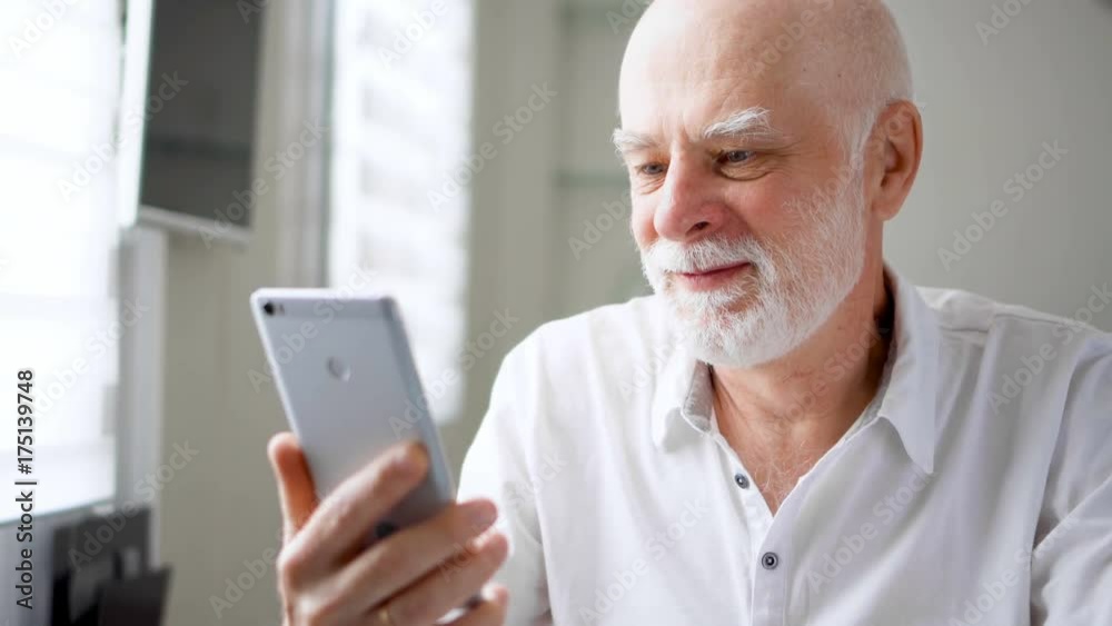 Senior man sitting at home with smartphone. Using mobile talking via messenger app Skype. Smiling waving hand in greeting. Remote freelance work on retirement, active modern lifestyle of older people Senior man sitting at home with smartphone. Using mobile talking via messenger app Skype. Smiling waving hand in greeting. Remote freelance work on retirement, active modern lifestyle of older people