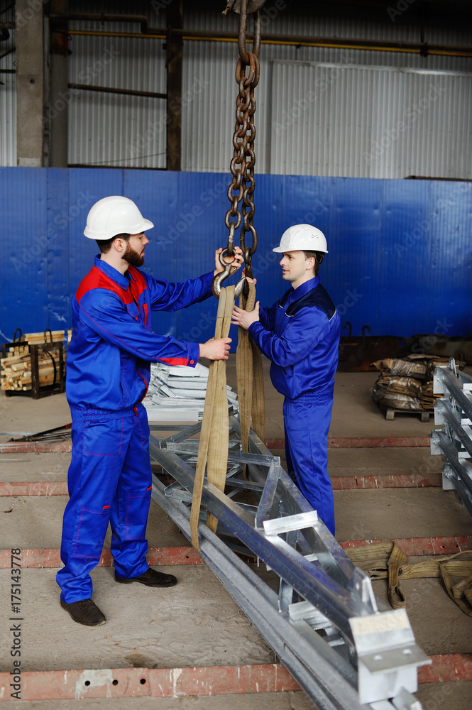 Two workers in white construction helmets load industrial products on the hook of the crane against the background of production Two workers in white construction helmets load industrial products on the hook of the crane against the background of production