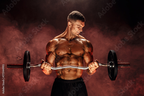 Studio portrait of topless bodybuilder performing biceps exercise with concentrated face over black background with smoke. Cutout. Very brawny guy bodybuilder.