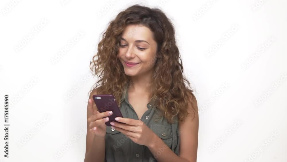 portrait of young caucasian woman with brown curly hair using mobile phone reading pleasant text message receiving good news isolated on white background. Concept of emotions portrait of young caucasian woman with brown curly hair using mobile phone reading pleasant text message receiving good news isolated on white background. Concept of emotions