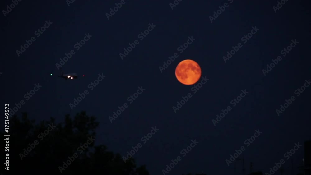 Wide, airplane flies by orange moon in night sky Wide, airplane flies by orange moon in night sky