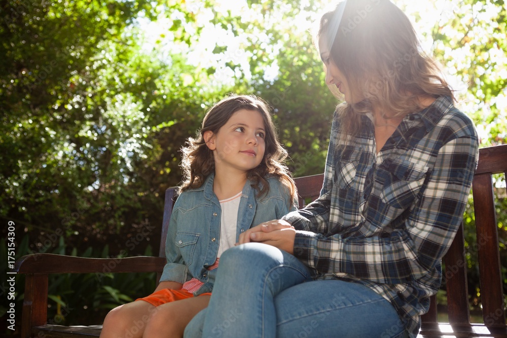 Fototapeta premium Girl sitting with mother on wooden bench against trees during