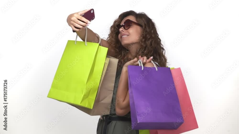 wonderful curly lady in sunglasses smiling making selfie photos holding a lot of packages with purchases after shopping isolated over white slomo. Concept of emotions