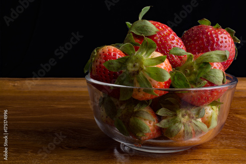 Strawberries in glass bowl on wooden tray against black background.