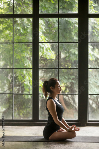 Free Download Young woman practicing yoga, sitting in Padmasana exercise, Lotus pose, mudra gesture, her eyes closed, working out, wearing sportswear, black shorts and top, indoor full length, window background - GFXLoad