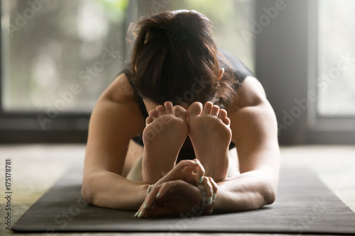 Young woman practicing yoga at home, stretching in Seated forward bend exercise, paschimottanasana pose, working out wearing black sportswear, indoor close up image studio background. Wellness concept
