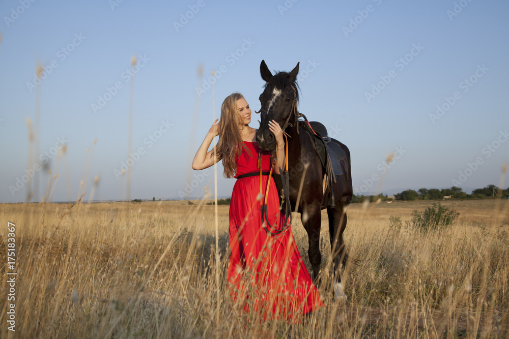 Beautiful blonde in red dress hugging a horse in field on background of countryside