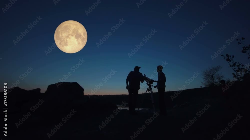 Black silhouettes of people looking through telescope on big red moon in dark twilight on shore.