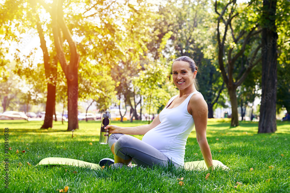 Pregnant woman holding bottle of water, sitting on yoga mat in park Pregnant woman holding bottle of water, sitting on yoga mat in park