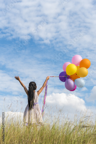 Happy girl holding colorful of air balloons on a green meadow with cloudy and blue sky