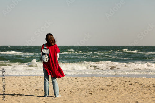 Young woman (brunette) in a red cardigan and light blue jeans, with a backpack, walks along the beach.