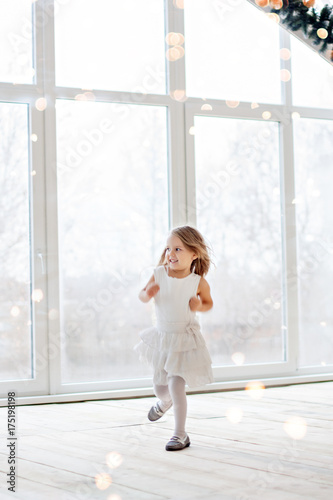 Little happy girl in a festive white dress, looks out the panoramic loft window, dances, decorates a Christmas tree in a pot with a garland of lights bokeh. Winter rest, waiting miracle of New Year