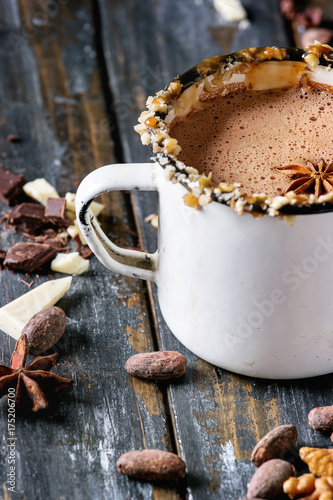 Vintage mug of hot chocolate, decor with nuts, caramel, spices. Ingredients above. Chopped dark and white chocolate, cocoa beans, anise over old wooden table. Dark rustic style. Close up