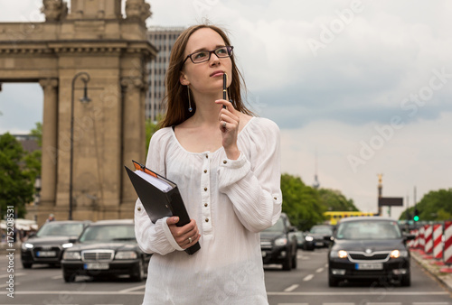 a young woman in glasses holding a pen and a folder and standing on the street; thoughts and ideas 