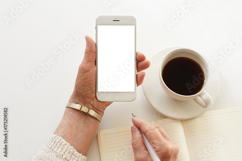 Mature businesswoman handwriting in copybook using mobile with blank white copy space display for your information. Close up of senior woman's hands holding cell phone; mug and notebook on desk