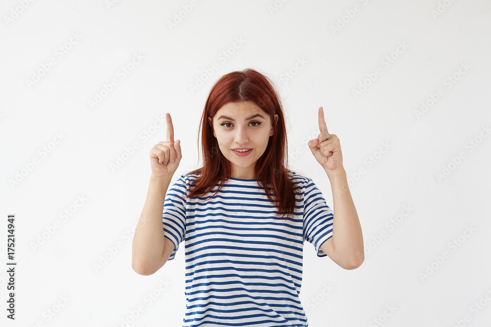 Isolated shot of attractive young European woman wearing stylish sailor shirt smiling and pointing both index fingers upwards, indicating something interesting on blank studio wall above her head Isolated shot of attractive young European woman wearing stylish sailor shirt smiling and pointing both index fingers upwards, indicating something interesting on blank studio wall above her head