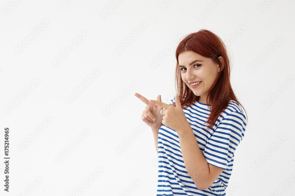 Just look at that! Pretty female with charming playful smile posing against white studio wall background, grinning broadly at camera and pointing fingers at copy space for your advertising content Just look at that! Pretty female with charming playful smile posing against white studio wall background, grinning broadly at camera and pointing fingers at copy space for your advertising content