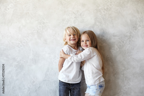 Horizontal picture of cute European little couple cuddling indoors: smiling beautiful girl hugging blonde handsome boy who feeling shy. Male and female kids wearing similar clothes, playing together