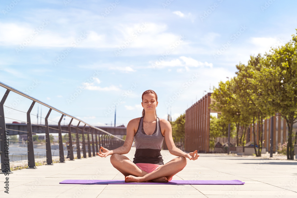 Young woman doing yoga lotus position in the city by the river, in sport clothes, relaxed and calm positive meditation.