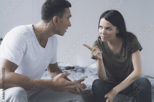 The woman angrily points a finger at the man who sits next to her on the bed and tries to justify himself