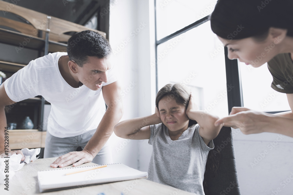 Obraz premium A man and a woman angrily scream at a small dark-haired girl who sits at a desk near the window, her hands over her ears