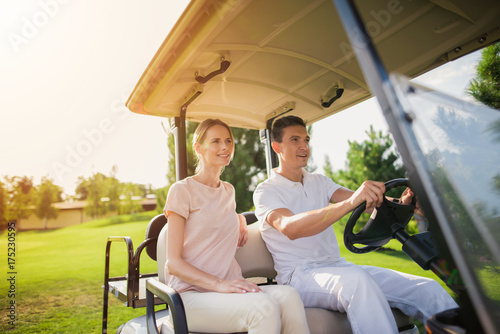 A couple is going to play golf. A man sits behind the wheel of a white golf car, a woman sits near