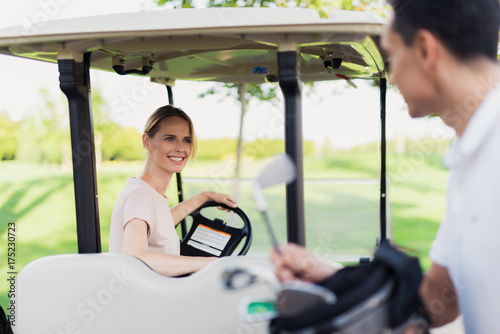 A man pulls out a golf club from the bag, a woman sits behind the wheel of a golf car