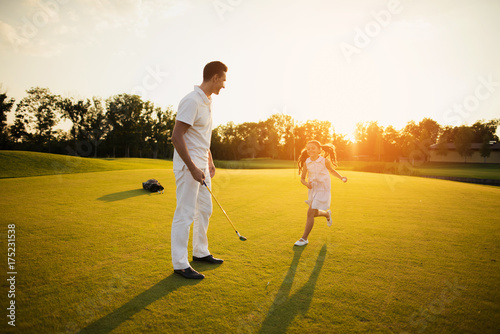 A girl with a golf ball in her hands runs to the man who stands near the golf club